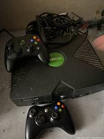 Photo showing original Xbox console with green circular Xbox logo, two black wired controllers with colored buttons, and a bundle of cables including power and video connectors.