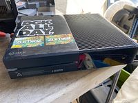 Image shows a black Xbox One console placed on a table. The console features multiple stickers on top including DGK ALL DAY and two Zumiez stickers. HDMI port is visible on the front. The console has some dust and signs of wear.