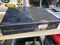 Black Xbox One console unit on a table, showing top side with scratches, tape labeled '303' on front right, disc drive slot, and vent grooves on the right side.