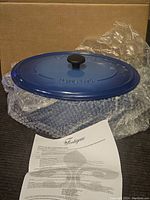 Blue enamel cast iron oval Dutch oven with lid wrapped partially in bubble wrap, alongside printed care instructions.