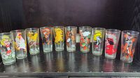 Set of 10 vintage 1970s Pepsi character glasses arranged in a row, showing colorful cartoon prints on clear glass.