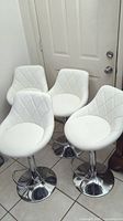 Four white adjustable height bar stools with diamond quilted backs arranged in a room corner.