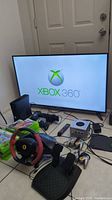 All items arranged with Sceptre TV displaying Xbox 360 logo, Xbox console, controller, Kinect, racing wheel, pedals, and stack of Xbox game cases