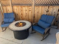 Wide view of two wicker rocking chairs flanking round fire pit table on concrete pad by wooden fence