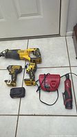 Photo showing DeWalt cordless reciprocating saw, two DeWalt cordless drills/drivers, Milwaukee M12 ratchet with battery and charger, and a single DeWalt 20V battery. Tools show light wear and dust.