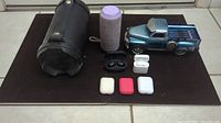 All items arranged on surface: three Bluetooth speakers - black cylindrical, gray fabric-wrapped cylindrical, blue truck style; four wireless earbud pairs with cases in front.