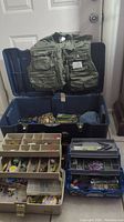 Fishing vest and three large tackle boxes partially open showing contents inside including lures and fishing line.