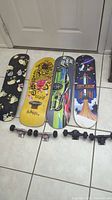 Four skateboard decks lined up on tile floor with attached trucks and wheels. Varied colorful graphics on decks. Visible wear and signs of use.