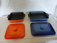 Two square casserole dishes with lids, one orange set and one blue set shown from above on a white fabric surface.