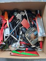 Top view of a cardboard box filled with assorted kitchen utensils including knives, ladles, spatulas, graters, and other tools in red, black, orange, and metallic colors.