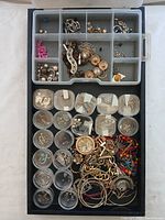 A top-view photo showing a plastic compartmentalized box holding an assortment of jewelry items like chains, earrings, pendants, and pink beads in organized sections.