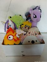 Assorted plush toys viewed from the front inside a white box, showing variety including a purple unicorn, green crocodile, orange chicken with glasses, character with yellow hair, and a koala.