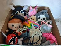 Box containing assorted plush toys and two small kids bags. Various characters including moose, girl, rainbow star, teddy bear, skull-faced and unicorn plushes.