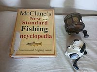 Photo showing McClane's New Standard Fishing Encyclopedia book with two fishing reels beside it on a light surface.