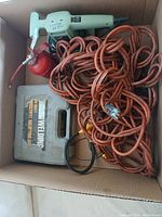 Top view of light green handheld electric sander, large coil of orange electrical cord, red metal oil can, and gray plastic case for soldering kit