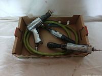 Three Mac air tools in a cardboard box including an impact wrench, a ratchet wrench, and a die grinder with a green air hose.