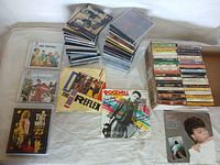 Overview of the lot showing stacks of single CDs, cassette tapes, and two vinyl records on a white surface.