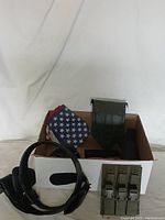 Box containing folded American flag, green military helmet carrier, magazine pouches, and black leather strap.