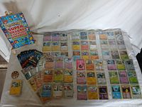 Pokémon cards arranged in nine clear plastic protective pages, one small container with loose cards, a plastic coin, and unopened booster packs displayed together with a Pokémon handbook