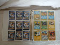 Binder open showing pages of Pokémon cards arranged in plastic sleeves. Card backs visible on one page, fronts of assorted cards visible on opposing page.