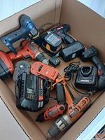 Box containing assortment of various used cordless drills, battery packs, battery chargers, and drill accessories.