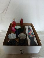 Box containing two lava lamps (one bottle shaped with pink liquid and one conical shaded with red-yellow-green gradient), two black lamp bases, and a boxed lava lamp accessory.