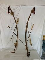 Photo showing two Native American style bows with arrows, fur, and feather decorations hung against a white backdrop.