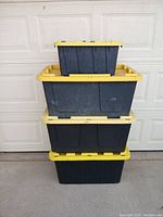 Stack of four plastic storage containers with yellow lids against a garage door, showing size difference between one small and three large containers.