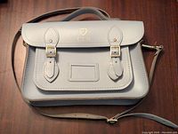 Light blue leather Cambridge Satchel displayed flat on wooden surface, showing front view with foldover flap, two gold buckles, outside pocket, adjustable crossbody strap, and gold initials 'CM' on flap.