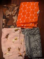 Four scarves folded and displayed: brown camouflage, orange with white dots, pink with gold cats, grey leopard print.