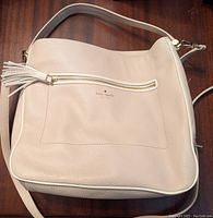 Front view of light tan textured leather Kate Spade handbag with white piping and tassel detail on front zipper pocket.