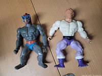 Two Masters of the Universe figures laid flat on a wooden surface showing front view. Beast Man figure on left, He-Man figure on right.