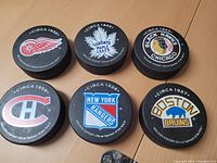 Top view of six hockey pucks each featuring a different Original Six NHL team logo (Detroit Red Wings, Toronto Maple Leafs, Chicago Blackhawks, Montreal Canadiens, New York Rangers, Boston Bruins) along with corresponding circa year.