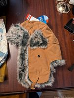 Light brown women's winter hat with gray and black faux fur lining visible around the edges and ear flaps. Hat has snap button on the side and chin strap for fastening.