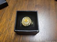 Gold-tone men's ring with Boston Bruins 1929 Stanley Cup emblem on top, set in black presentation box on wooden surface.