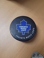 Top side of hockey puck with Toronto Maple Leafs blue and white logo on black background.