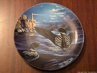 Decorative plate featuring two loons on a moonlit lake with lighthouse in background, signed by artist Jim Hansel.