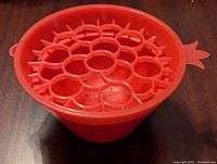Red plastic pomegranate seed remover tool showing interior grid insert inside bowl with pomegranate-seed shaped cutouts and side tab handle
