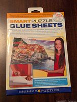 Front view of Smart Puzzle Glue Sheets box showing product name, seaside puzzle image, and model with completed puzzle.