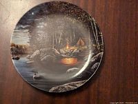 Front view of decorative plate showing nighttime lakeside camping scene with campfire, tent, trees, rocks, and a loon swimming on the lake.