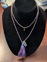Front view of the two-strand purple beaded necklace displayed on a black velvet bust, showing the tassel and charm details with tag attached.