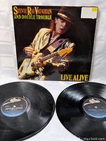 Front of album cover with Stevie Ray Vaughan in gold jacket playing guitar, two vinyl records in foreground