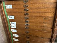 Wooden cabinet with multiple labeled drawers; one drawer open revealing plastic trays of watch parts