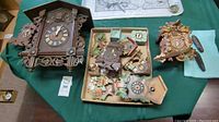Multiple wooden cuckoo clocks and box of assorted parts displayed on a green surface