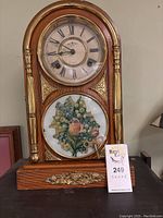 Front view of the vintage wooden mantel clock with gold decorative details, Roman numeral clock face, floral image below, and winding key leaning on the bottom glass panel.