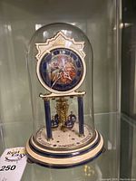 Front view of the vintage porcelain anniversary clock inside glass dome, showing detailed clock face with classical figure artwork, blue roman numerals, and gold accents.