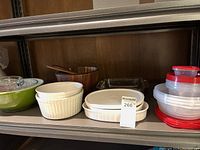 Shelf of kitchenware items including green Pyrex mixing bowl, white ceramic baking dishes, clear glass baking dishes, brown mixing bowls and plastic storage containers with lids.