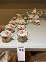 Photo showing six American Beauty tea cups with pink rose pattern, gold trim, and six matching saucers arranged on table alongside Lenora tea pot, cream jug, and sugar bowl with orange and purple floral patterns.