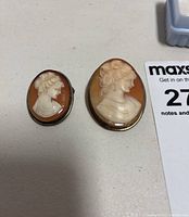 Two cameo brooches side by side on a flat surface, showing front view of carved female profiles. Larger and smaller ovals shown.
