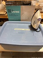 Blue plastic storage bin labeled 'Emergency Supplies' with Black & Decker iron placed on top and 12 inch for Living oscillating desk fan box behind.